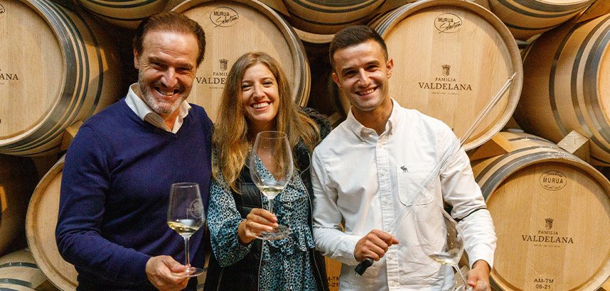 Drei Personen mit Weingläsern vor Fässern der Weinkellerei Valdelana.