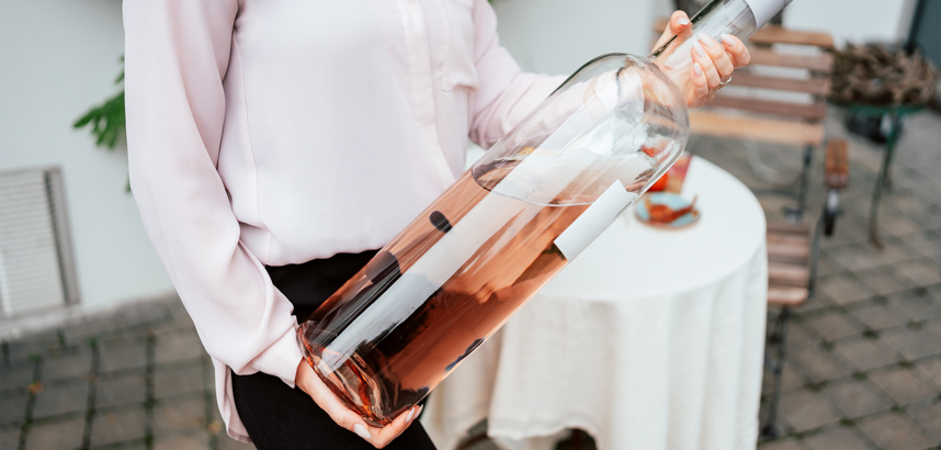 Person hält riesige Flasche Roséwein im Aussenbereich nahe Tisch.